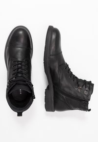 Svarta läderstövlar har en rund tå, snörning framtill och sidokomprimerade blixtlåshål. De har en strukturerad gummisula och en slät yta.