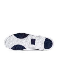 Semelle de sneaker blanche avec motifs circulaires bleus antidérapants au talon, à la plante du pied et au milieu, montrant une bande de roulement en zigzag texturée et une étiquette de marque au centre.