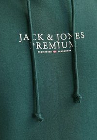 Dunkelgrüne Kapuzenjacke aus weichem Material mit einer Kängurutasche, Kordelzügen und dem weißen Aufdruck "JACK & JONES PREMIUM".
