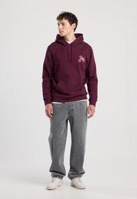 Burgunderroter Kapuzenpullover mit Fronttasche und Logodetail. Kombiniert mit grauen, locker sitzenden Jeans und weißen Turnschuhen. Glatte Stofftextur.