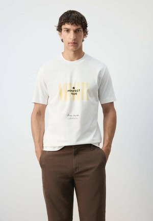 Joven con camiseta blanca con rayas amarillas y texto "Perfect Sun", combinado con pantalones marrones, de pie con las manos en los bolsillos.