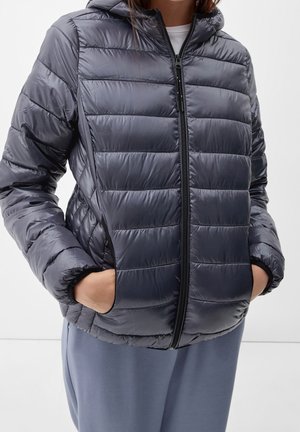 Person trägt eine glänzende dunkelgraue gesteppte Kapuzenjacke mit Reißverschluss und den Händen in den Taschen, kombiniert mit lockeren grauen Hosen.