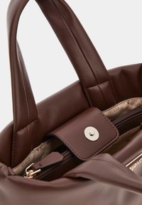 Braune Lederhandtasche mit glatter Oberfläche, Griffen oben und einem Verschluss mit goldfarbener Druckknopf. Das Innere verfügt über eine Reißverschlusstasche und eine gefütterte Innenverkleidung mit Markenlogo.