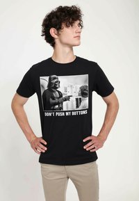 Černé bavlněné tričko s potiskem černobílého grafického zobrazení interakce postav, s textem "NEPŘIDÁVEJ MI NA STAROSTECH" pod tím.