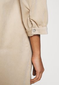 Bras d’une personne portant une chemise beige à manches longues avec un poignet remonté et maintenu par un bouton.