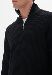 Suéter negro con media cremallera y un patrón de ribetes texturizados, que cuenta con una cremallera plateada y un cuello alto. La tela parece cálida y duradera.