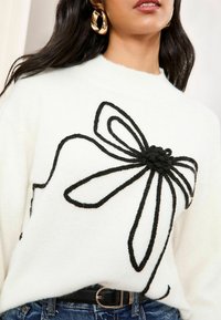 Weißer gestrickter Pullover mit einem großen schwarzen Schleifendessin. Die Textur ist weich, mit einer lässigen Passform und einem Rundhalsausschnitt.