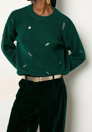 Vrouw draagt een donkergroene trui met gouden geborduurde harten en woorden, gecombineerd met een donkere broek en een metalen riem.