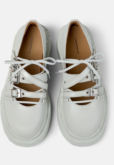 Para białych skórzanych butów z grubymi podeszwami, krzyżującymi się paskami ze srebrnymi klamrami i białymi sznurowadłami zawiązanymi na kokardki, z marką CamperLab wewnątrz.