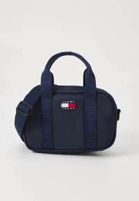 Tmavě modrá látková kabelka s dvojitými rukojeťmi a odnímatelným popruhem, zdobená nášivkou s logem a texturovanými bočními akcenty. Zapínání na zip.