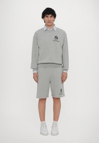 Set cu hanorac gri și pantaloni scurți, confecționat dintr-un material moale, având un logo negru pe ambele piese. Modelul poartă adidași albi și o cămașă cu guler de culoare albastru deschis.