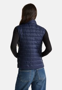 Žena s dlouhými tmavými vlasy, oblečená v námořnické prošívané vestě přes černý dlouhý rukáv a modré džíny, stojí zády k fotoaparátu.