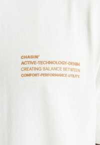 Bijela tkanina s rebrastom teksturom na kojoj je narančastim slovima otisnut tekst: "CHASIN' ACTIVE-TECHNOLOGY-DENIM STVARANJE BALANSA IZMEĐU UDOBNOSTI-PERFORMANSE-KORISNOSTI."