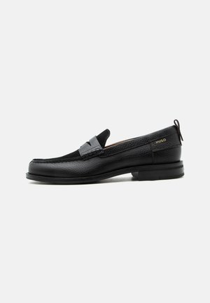 Mocassin en cuir noir avec détail de bride grise, talon bas et semelle texturée, marqué "HUGO" en or sur le côté.