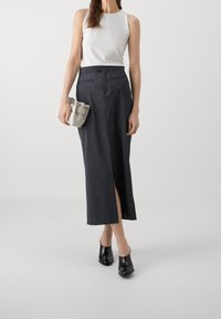 Grå, halvlang kjol med framslits, paired med en vit ärmlös topp. Modellen håller en liten, ljusgrå väska. Svarta, stängda skor.