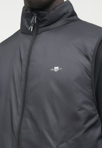 Schwarze, leichte Weste mit hohem Kragen, Reißverschluss und dezentem GANT-Logo. Glatte, matte Textur, entworfen zum Tragen über einem Hemd.