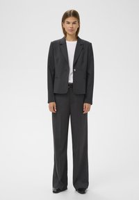 Szürke szabott blézer egy gombbal, laza szürke nadrággal párosítva. Alatta egy fehér póló látható. Simító anyag és strukturált fazon.