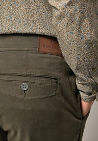 Khakihosen mit glatter Textur, ausgestattet mit einer einzelnen Gesäßtasche und einem braunen Lederetikett mit der Aufschrift "BOSTON PARK." Das Hemd hat ein Blattmuster.