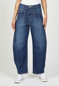 Blaue Denim-Hose mit weitem Bein und hoher Taille, zwei frontalen Taschen und sichtbaren Nähdetails. Kombiniert mit weißen Sneakers für einen Kontrast.