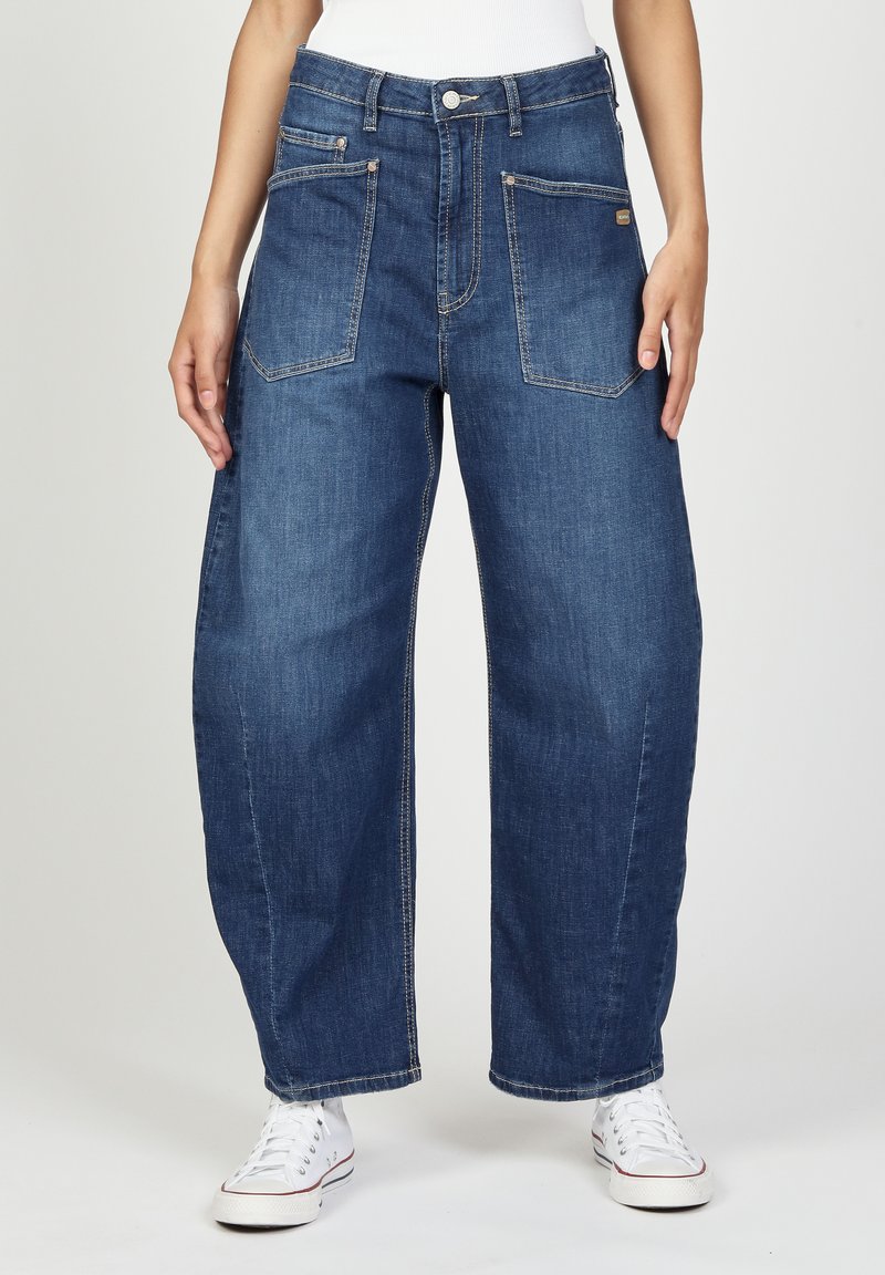 Blaue Denim-Hose mit weitem Bein und hoher Taille, zwei frontalen Taschen und sichtbaren Nähdetails. Kombiniert mit weißen Sneakers für einen Kontrast.