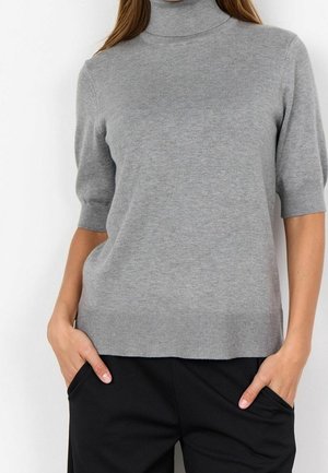 Femme portant un pull à manches courtes col roulé gris clair avec un ourlet côtelé et un pantalon noir, les mains dans les poches.