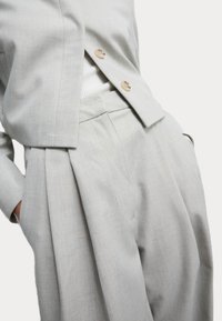 Hellgrauer, taillierter Blazer mit zwei Knöpfen, kombiniert mit hochgeschnittenen, gefalteten Hose. Aus strukturiertem Stoff gefertigt, der klare Linien und Taschen präsentiert.
