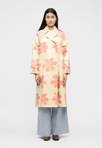 Γυναίκα που φοράει μακρύ κρεμ παλτό με μεγάλα ρόδινα λουλουδάτα σχέδια, φαρδιά ανοιχτά μπλε παντελόνια και καφέ παπούτσια, στέκεται σε απλό φόντο.