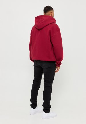 Mężczyzna w czerwonej, oversize'owej bluzie z kapturem, czarnych obcisłych dżinsach i białych tenisówkach, stojący tyłem na tle jednolitego białego tła.