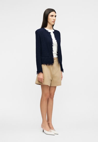 Γυναίκα που στέκεται φορώντας μπλε ναυτικό υφασμάτινο σακάκι με ανοιχτό μπροστινό μέρος, λευκό τοπ, μπεζ σορτς και λευκές μυτερές γόβες σε απλό φόντο.