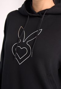 Hanorac negru cu un design alb în contur al unui iepure cu o formă de inimă. Are un buzunar tip cangur și un glugă cu șnur.