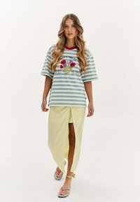 Pruhované oversized tričko ve světle zelené a bílé barvě s květinovou grafikou; spárované se světle žlutou sukní s předním rozparkem.