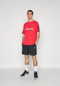 Punainen urheilupaita, jossa on mustat raidat hihoissa ja "ADIDAS SPORTSWEAR" -logo. Mustat shortsit ja mustat urheilukengät. Valkoiset sukat.
