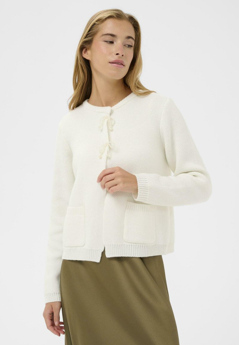 Cardigan blanc en tricot avec un col rond, fermeture à nouer sur le devant et deux poches plaquées, présentant un tissu texturé et des poignets côtelés.