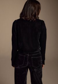 Zwart fluwelen top met lange mouwen, voorzien van subtiele plooien bij de schouders. Gecombineerd met bijpassende zwarte fluwelen broek, beide met een gladde textuur.