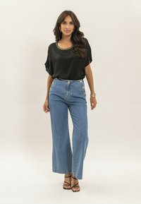 Blouse en satin noir avec des bordures dorées, jeans large en denim bleu clair taille haute avec des accents de boutons, et sandales noires à lanières.