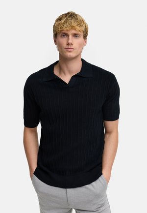 Blonder Mann mit kurzen Haaren, der einen schwarzen kurzärmeligen Pullover mit Kragen und hellgraue Hosen trägt und die Hände in den Taschen hat.