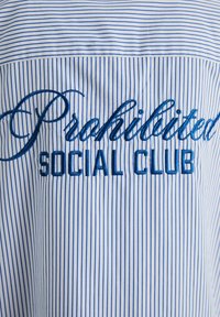 Prúžkovaná košeľa v bielej a modrej farbe s kurzívou napísaným "Zakázané" a tučným "SOCIAL CLUB" vyšitém kontrastnou modrou niťou na prednej strane.