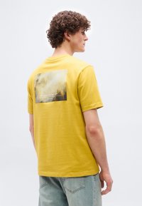 Νεαρός άνδρας με σγουρά μαλλιά που φοράει κίτρινη μπλούζα με γραφικό ομίχλης δάσους και κείμενο στην πλάτη, συνδυασμένη με ανοιχτόχρωμο μπλε τζιν.