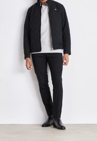 Black quilted jacket with a high collar, layered over a gray sweater, paired with black trousers and sleek black boots. Minimalist design.