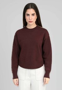 Pull en tricot bordeaux avec un col rond côtelé et des poignets. Présente des surpiqûres décoratives blanches le long des manches.
