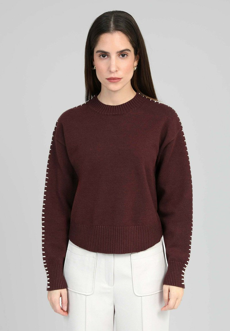 Pull en tricot bordeaux avec un col rond côtelé et des poignets. Présente des surpiqûres décoratives blanches le long des manches.