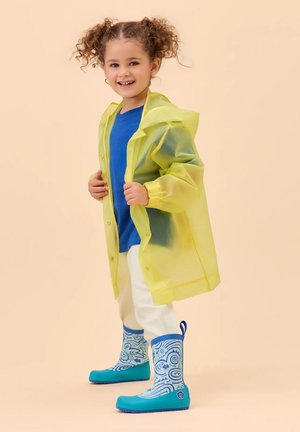 Lächendes Kind mit lockigem Haar, das einen gelben Regenmantel, ein blaues Hemd, weiße Hosen und blaue, gemusterte Gummistiefel trägt und vor beigem Hintergrund steht.