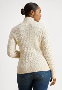 Moteris su tamsiais plaukais surištais į kuoduką, dėvinti auksinius auskarus, kreminio atspalvio megztinį su raišteliu ir aukštu apykakle, bei mėlynas džinsus, žvelgianti šonu prieš balta foną.