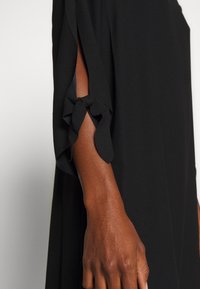 Czarna sukienka z krótkimi rękawami, z detalem w postaci wiązania przy łokciu. Materiał jest gładki i zwiewny, z delikatnym połyskiem.