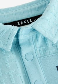 Camisa de polo texturizada azul claro con una placket de botones, que presenta patrones geométricos en relieve y una etiqueta negra que dice "BAKER by TED BAKER."
