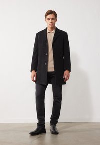 Abrigo de lana negro con cuello muesca, dos botones y bolsillos laterales. Combinado con un polo beige, jeans oscuros ajustados y botas negras.