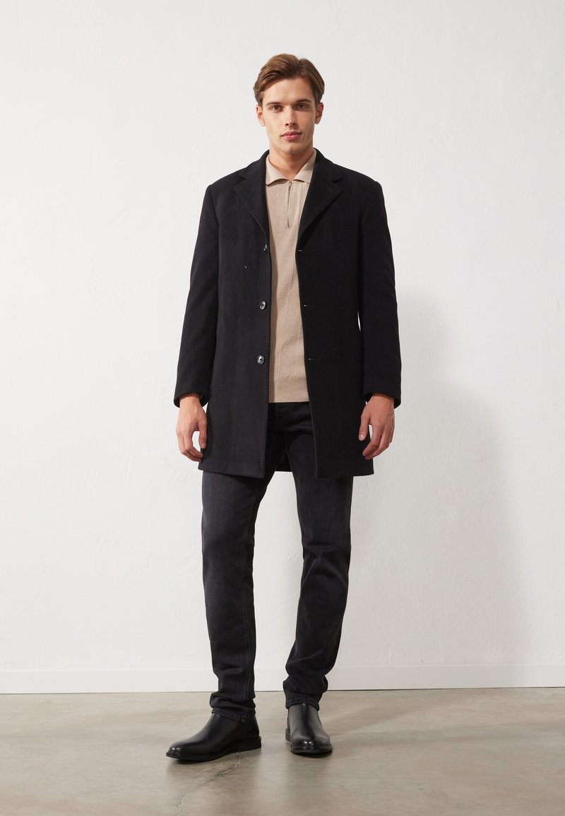 Abrigo de lana negro con cuello muesca, dos botones y bolsillos laterales. Combinado con un polo beige, jeans oscuros ajustados y botas negras.