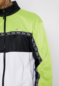 Chaqueta con bloques de color en verde, negro y blanco. Presenta un cierre de cremallera, cuello alzado y logo de Fila en rojo. Material suave.