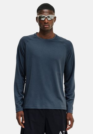 Mand iført mørkeblå, langærmet tekstureret skjorte, sorte shorts og reflekterende hvide solbriller, stående foran en ensfarvet lys baggrund.