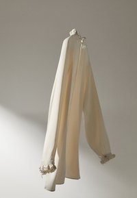 Blouse couleur crème à manches longues. Présente un col et des poignets volantés, fabriquée dans un tissu lisse et léger. Texture douce, design minimaliste.